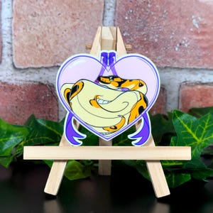 May include: A sticker of a yellow and black snake curled up inside a pink heart with purple ribbon accents.