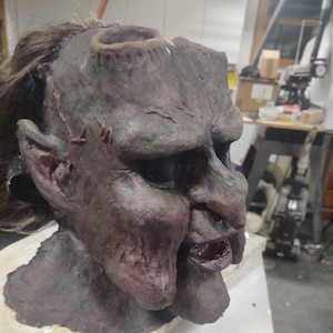 May include: A detailed, dark-colored mask with a grotesque, weathered appearance. The mask features a large nose, open mouth, and deep-set eyes, with visible sutures and a hole in the head. The mask is likely for Halloween or theatrical use.
