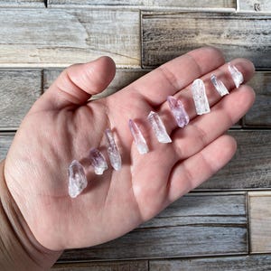 May include: A collection of small, clear quartz crystals with hints of purple, arranged in a hand. The crystals vary in size and shape, with pointed tips. The background is a wooden surface.