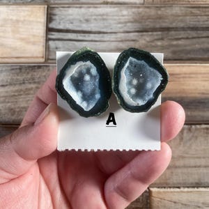 May include: Two geode earrings with a dark green border and a white and blue crystal center. The earrings are displayed on a white card with the letter "A" printed on it. The background is a wooden surface.