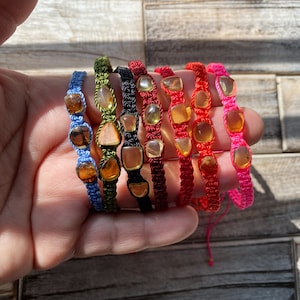 May include: Eight colorful braided bracelets with amber gemstone beads. The bracelets are in shades of blue, green, black, red, and pink.