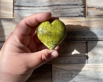 May include: A green heart-shaped stone with a marbled pattern. The stone is being held in a hand.