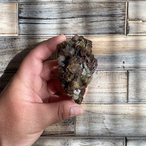 May include: A cluster of raw, uncut crystals in shades of brown, tan, and clear. The crystals have a rough, textured surface and are held in a hand against a wooden background. The mineral specimen appears to be a natural, unpolished stone.