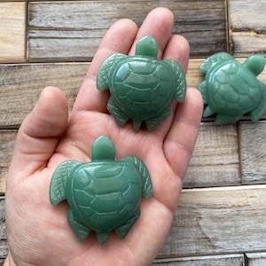 May include: Three carved green turtle figurines. The turtles have detailed textured shells, flippers, and heads. The figurines are a vibrant green colour and appear to be made of polished stone. The turtles are displayed on a wooden surface.