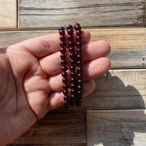 May include: Three strands of dark red beaded bracelets. Each bracelet is made of small, round beads strung together. The bracelets are held against a wooden background, with the sunlight casting shadows.