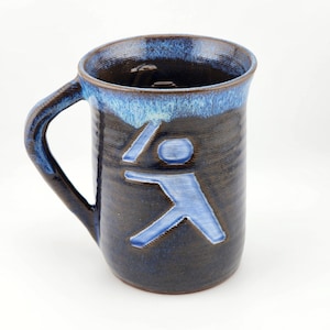 May include: A handmade ceramic mug with a dark brown body and a blue rim. The mug features a blue abstract figure design. The mug has a large handle and is suitable for hot or cold beverages.