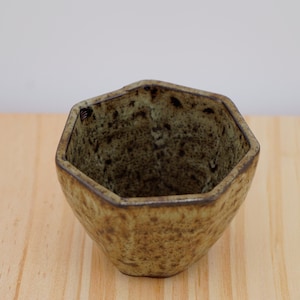 May include: A hexagonal ceramic bowl with a speckled, earthy brown and green glaze. The bowl has a slightly rough texture and sits on a light-colored wooden surface. The interior of the bowl is a darker shade of green.