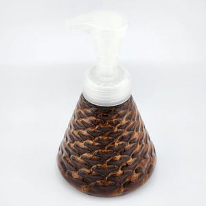 May include: A brown ceramic soap dispenser with a textured, honeycomb-like pattern. The dispenser has a white plastic pump and a conical shape, suitable for a bathroom or kitchen. The dispenser is designed to hold liquid soap.