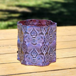 May include: A handcrafted ceramic vase with a textured surface and a unique pattern. The vase features a blend of purple and blue hues, with a reddish-brown interior. The hexagonal shape adds a modern touch to this decorative piece.
