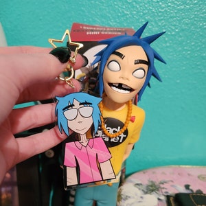 May include: A clear acrylic keychain featuring a cartoon illustration of a blue-haired person wearing a pink shirt and blue jeans. The keychain is attached to a gold star charm.