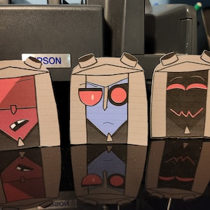 May include: Three paper cut-outs of cartoon characters with different facial expressions. The characters are wearing brown hooded cloaks with black collars. The characters have red, blue, and black faces with different facial features.