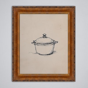 Op de afbeelding: Een ingelijste pen- en inkttekening van een afgedekte braadpan. Het kunstwerk zit in een goudkleurige lijst. De tekening is een eenvoudige lijntekening van de pan, met een deksel en handvatten. De achtergrond is lichtbeige.