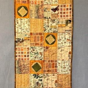 Op de afbeelding: Een gewatteerde wandhanger met een patchwork-ontwerp. De quilt heeft vierkanten met verschillende patronen, waaronder bloemenprints, kaarten en vintage efemera. Groene ruitvormen zijn in het ontwerp genaaid. De rand is donkerbruin.