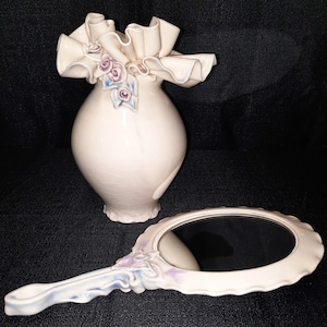 May include: A white ceramic vase with a ruffled top and floral accents, paired with a white ceramic hand mirror with a floral design.