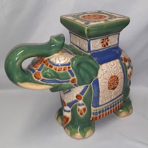 Może przedstawiać: Ceramiczny stołek ogrodowy w kształcie słonia w odcieniach zieleni, niebieskiego, bieli i brązu. Słoń ma kwadratowy blat i jest ozdobiony skomplikowanymi wzorami i wzorami. Trąba jest zakręcona, a stołek ma szczegółowe, teksturowane wykończenie.