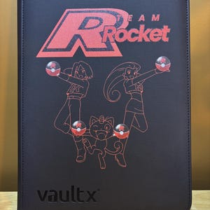 May include: A black zippered card binder featuring a red Team Rocket logo and line art of Jessie, James, and Meowth holding Poké Balls. The front cover displays the text Team Rocket in red and the brand name vaultx embossed in the bottom corner.