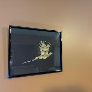 May include: Framed artwork featuring a bird silhouette crafted from golden bullet casings against a dark background. The black frame contrasts with the warm tones of the wall, creating a striking visual. This piece is a unique decorative item.