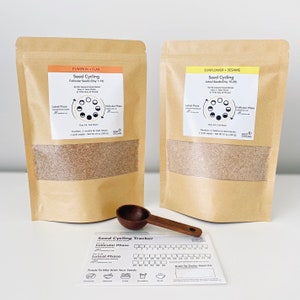 May include: Two brown paper bags filled with seeds, labelled "Pumpkin + Flax" and "Sunflower + Sesame". The bags are next to a wooden scoop and a printed card with a seed cycling tracker, showing the follicular and luteal phases of the menstrual cycle.
