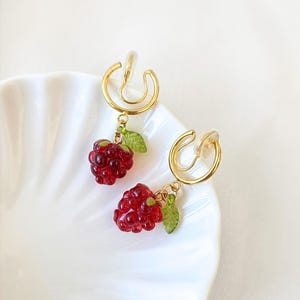 Pode incluir: Um par de brincos de press&atilde;o com um design em metal dourado. Cada brinco tem um pingente de framboesa vermelha transl&uacute;cida com folhas verdes. Os brincos s&atilde;o exibidos em um prato branco com bordas onduladas. O texto "CLIP ON ATELIER" est&aacute; na parte inferior.