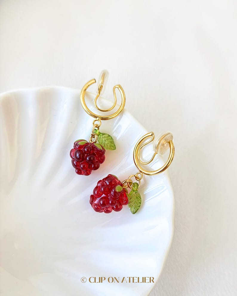 Pode incluir: Um par de brincos de press&atilde;o com um design em metal dourado. Cada brinco tem um pingente de framboesa vermelha transl&uacute;cida com folhas verdes. Os brincos s&atilde;o exibidos em um prato branco com bordas onduladas. O texto "CLIP ON ATELIER" est&aacute; na parte inferior.