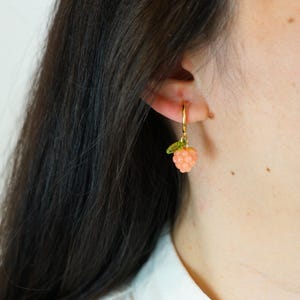 Pode incluir: Brinco de argola dourado com um pingente de framboesa cor de p&ecirc;ssego e uma pequena folha verde. O brinco &eacute; usado na orelha. A imagem est&aacute; em um fundo branco. O texto "CLIP ON ATELIER" est&aacute; na parte inferior.