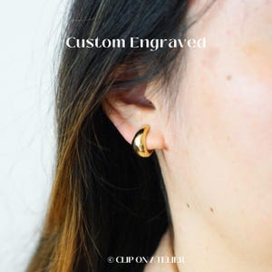 May include: Gold-colored, custom engraved hoop earrings. The earrings are a crescent shape and are worn on the ear. The text "Custom Engraved" is above the earrings. The brand name "CLIP ON ATELIER" is at the bottom.