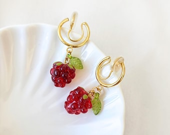 May include: A pair of gold-toned clip-on earrings featuring dangling red raspberry charms and small green leaves. The earrings have a circular gold metal hoop design with clear plastic cushions for comfort, showcasing a detailed fruit motif.