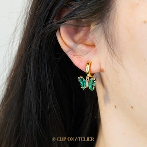 May include: Gold hoop earrings with a green butterfly pendant. The butterfly has a gemstone design and a small diamond accent. The earrings are shown on a person's ear.