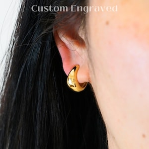 May include: A close-up of a gold-colored, chunky hoop earring. The earring is worn on an ear, with the text "Custom Engraved" at the top and "CLIP ON ATELIER" at the bottom. The earring has a smooth, polished surface.