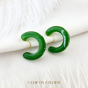 May include: A pair of green clip-on earrings with a curved, half-circle shape. The earrings are made of a glossy, opaque material and have a gold-toned clip.