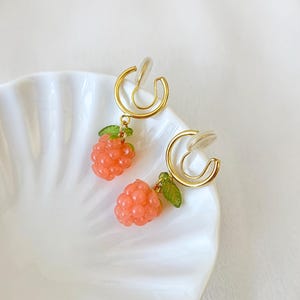 Pode incluir: Brincos de press&atilde;o com um design de argola dourada. Cada brinco tem um pequeno pingente de framboesa cor-de-coral com folhas verdes. Os brincos s&atilde;o exibidos em um prato branco com bordas onduladas. O texto "CLIP ON ATELIER" est&aacute; na parte inferior.