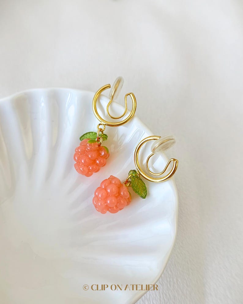 Pode incluir: Brincos de press&atilde;o com um design de argola dourada. Cada brinco tem um pequeno pingente de framboesa cor-de-coral com folhas verdes. Os brincos s&atilde;o exibidos em um prato branco com bordas onduladas. O texto "CLIP ON ATELIER" est&aacute; na parte inferior.