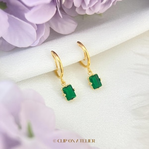 May include: A pair of gold hoop earrings featuring rectangular emerald green gemstone drops. Each earring consists of a thin gold hoop connected to a faceted green stone in a gold setting. The text CLIP ON ATELIER is visible at the bottom.