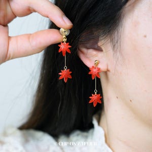 May include: Red maple leaf earrings with gold-colored accents. The earrings feature a clip-on design and a delicate chain suspending the maple leaf charms. The earrings are a vibrant red color and are shown on a person.
