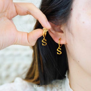 May include: Gold hoop earrings with a dangling letter 'S' charm. The earrings are shown on a person's ear.