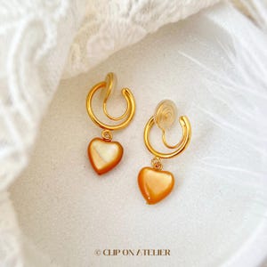 May include: A pair of gold-colored clip-on earrings. Each earring features a heart-shaped, orange-toned pendant suspended from a curved, gold-toned hoop. The earrings have a clip-on mechanism. The text "CLIP ON / ATELIER" is at the bottom.