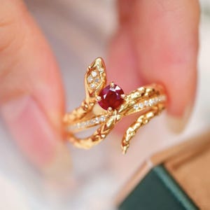 Puede incluir: Un anillo de serpiente de oro con una piedra preciosa de rubí rojo y diamantes más pequeños. El anillo está diseñado con dos cabezas de serpiente entrelazadas.