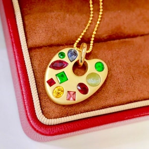 May include: A gold-colored pendant necklace featuring a palette design, adorned with various colorful gemstones. The pendant is suspended from a delicate gold chain. The necklace is presented in a red jewelry box with a brown interior.