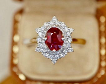 May include: A red ruby gemstone ring with a diamond halo setting. The ring is in a small, brown box.