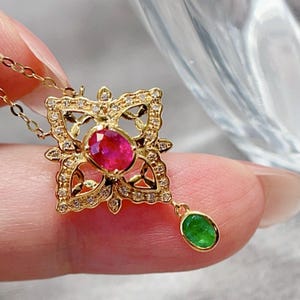 Puede incluir: Un collar de oro con un colgante que tiene una piedra preciosa de rubí roja engastada en un diseño de filigrana y una piedra preciosa de esmeralda verde colgante.