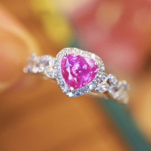 May include: A pink sapphire engagement ring with a heart-shaped halo setting. The ring is set in white gold and features a delicate, twisted band.