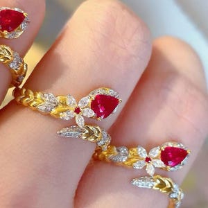 May include: Two gold rings with red ruby stones and white diamonds set in a floral design. The rings are on a finger.