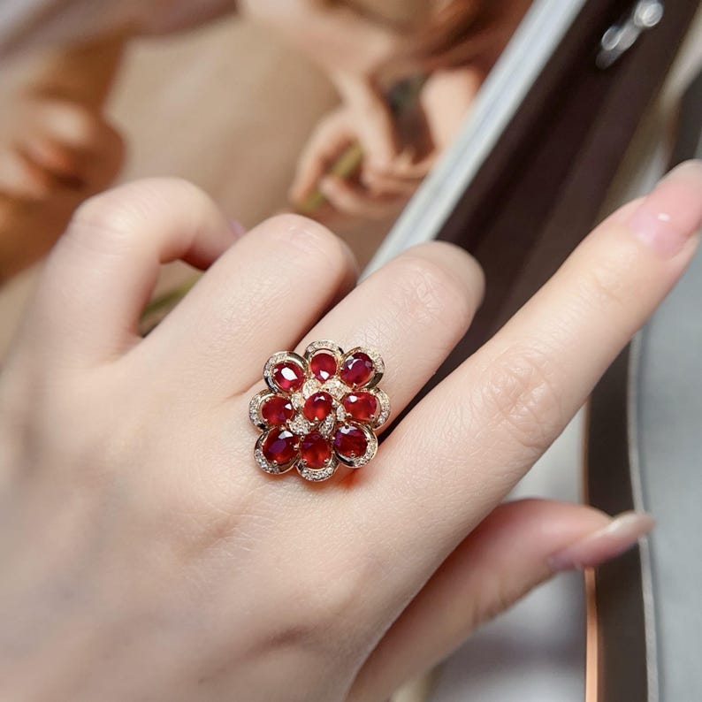 Puede incluir: Un anillo de oro con forma de flor con piedras preciosas rojas. Los p&eacute;talos del anillo est&aacute;n formados por piedras rojas, con peque&ntilde;as piedras transparentes alrededor. El anillo se lleva en un dedo.