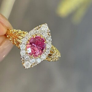 May include: A gold ring with a pink gemstone surrounded by a halo of smaller diamonds. The ring has a delicate, intricate design.