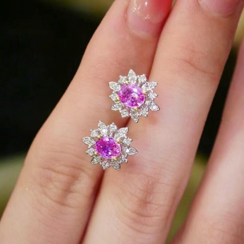May include: A pair of pink sapphire earrings with a floral design. Each earring features a pink oval-shaped sapphire surrounded by a halo of smaller diamonds.