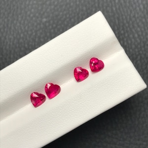 May include: Four heart-shaped gemstones, each a vibrant pink, are displayed on a white surface. The gemstones are arranged in a row, with the largest at the top and the smallest at the bottom.
