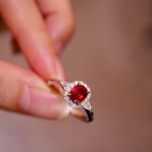 Könnte beinhalten: Ein silberner Ring mit einem kissenförmigen, tiefroten Edelstein, umgeben von einem Halo aus kleinen, klaren Steinen. Der Ring hat ein zartes Design mit zusätzlichen Akzentsteinen auf beiden Seiten des zentralen Edelsteins. Ein feines Schmuckstück.