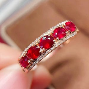 May include: A gold ring featuring a row of five oval-cut vibrant red rubies set horizontally. The central rubies are flanked on both the top and bottom edges by thin bands of small, clear, sparkling pavé-set stones, creating a layered design.