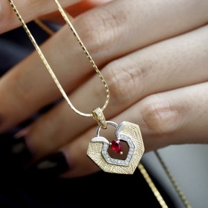 Könnte beinhalten: Eine Halskette aus Gold und Silber mit einem herzförmigen Anhänger, der mit einem roten Edelstein und kleinen klaren Edelsteinen besetzt ist.