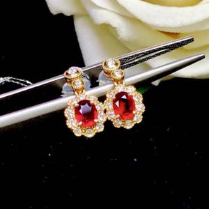 Puede incluir: Un par de pendientes de oro con piedras preciosas de rubí rojo rodeadas de diamantes más pequeños. Los pendientes tienen un diseño floral delicado.